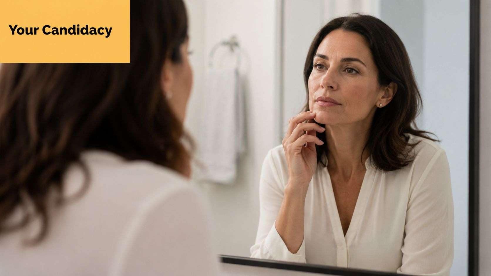 A middle aged woman looking in a bathroom mirror and touching her chin while considering aesthetic choices.